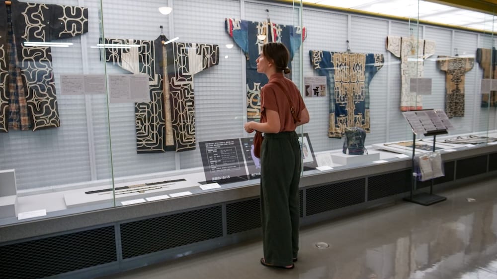 Audrey Bergner observa prendas tradicionales con diseños expuestas detrás de un vidrio dentro de Hakodate City Museum of Northern Peoples, en Hakodate, Hokkaido, Japón, con varias vestimentas alineadas en la galería.