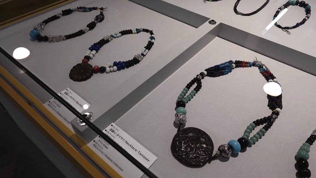 Glass-bead tamasay necklaces with round metal medallions in a display case at the Hakodate City Museum of Northern Peoples in Hakodate, Hokkaido, Japan, with bilingual labels naming the Kodama Collection.