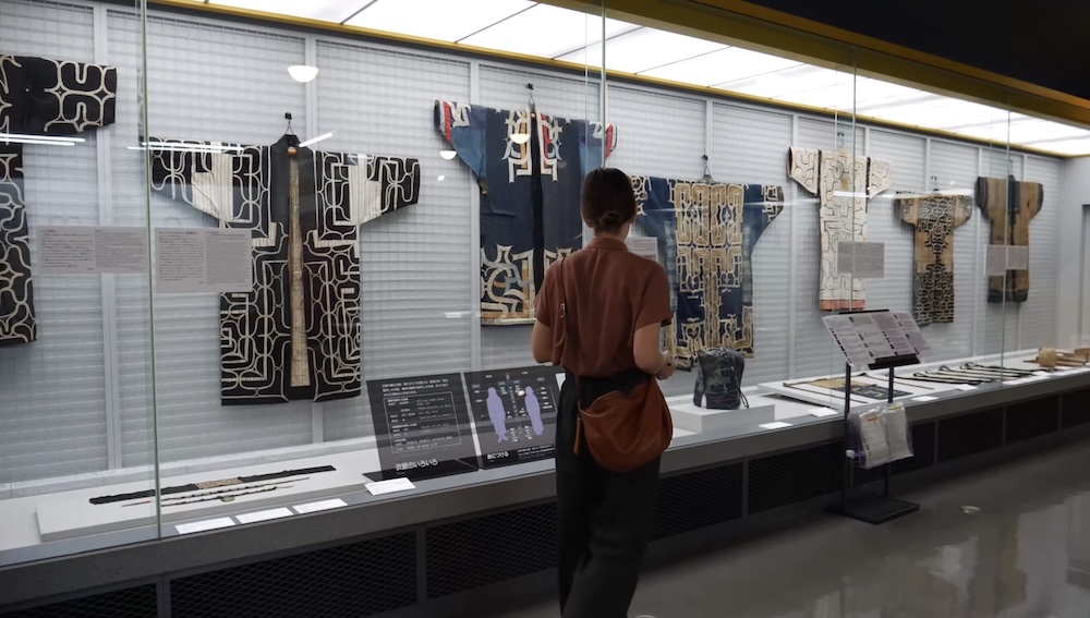 Audrey Bergner Viewing Traditional Ainu Clothing at Hakodate City Museum of Northern Peoples in Hokkaido Japan Audrey Bergner examining traditional Ainu robes with bold geometric patterns displayed behind glass at the Hakodate City Museum of Northern Peoples in Hokkaido, Japan, with interpretive panels and additional textiles visible.