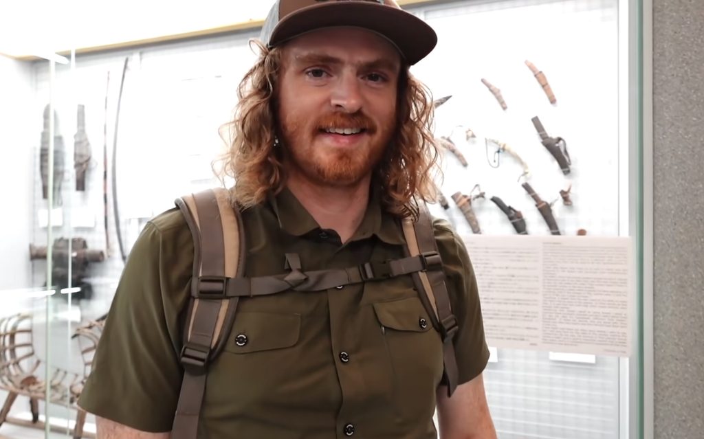 Samuel Jeffery in a brown cap, olive shirt and backpack, smiling in front of glass cases holding a bow, sheathed blades and a sled frame at the Hakodate City Museum of Northern Peoples in Hakodate, Hokkaido, Japan.