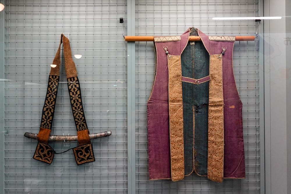 Glass display at the Hakodate City Museum of Northern Peoples in Hakodate, Hokkaido, Japan, showing a sheathed blade on an embroidered shoulder belt beside a sleeveless purple garment with gold brocade panels.
