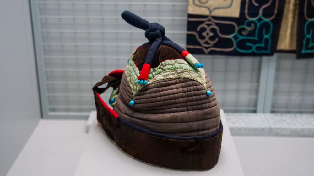 Quilted brown hat with a knotted navy top, red trim and blue beads on display at the Hakodate City Museum of Northern Peoples in Hakodate, Hokkaido, Japan, beside an embroidered robe.