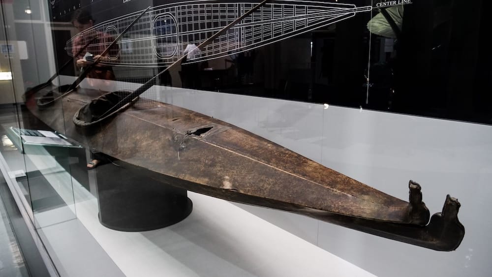 Long dark skin-covered boat with a forked bow and open cockpits in a glass display at the Hakodate City Museum of Northern Peoples in Hakodate, Hokkaido, Japan, in front of a line drawing marked Center Line.