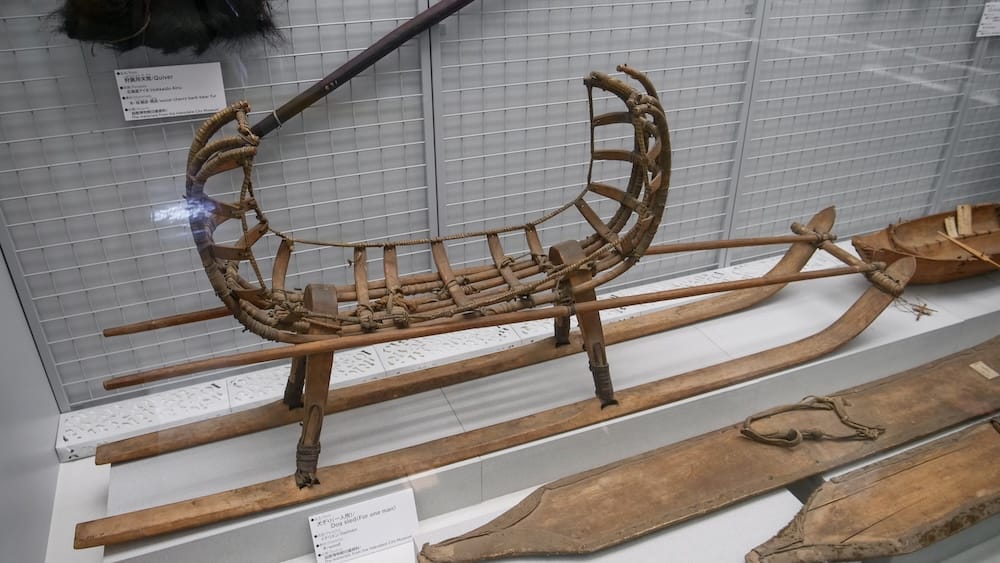 Wooden dog sled for one person with a curved lashed frame on long runners, shown behind glass at the Hakodate City Museum of Northern Peoples in Hakodate, Hokkaido, Japan, with a small boat at right.