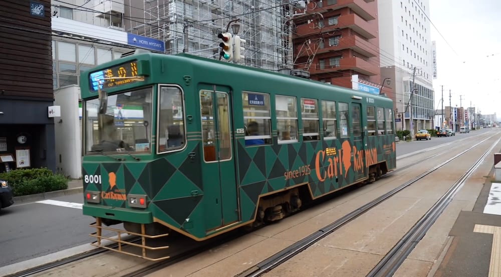 A dark green Hakodate city tram numbered 8001 with orange lettering runs along street tracks in Hakodate, Hokkaido, Japan, past a hotel sign, scaffolded building and traffic on an overcast day.