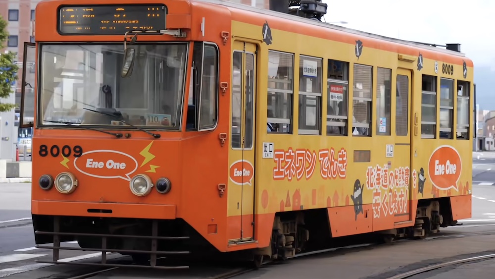 Hakodate City Tram Number 8009 in Hokkaido Japan Featuring Orange and Yellow Ene One Advertising Livery Orange and yellow Hakodate city tram number 8009 with Ene One advertising on its exterior, photographed on streetcar tracks in Hakodate, Hokkaido, Japan, showing its front windows, passenger doors and overhead electrical equipment.