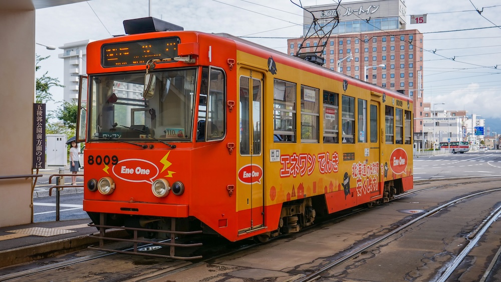 Orange and Yellow Hakodate City Tram Number 8009 on the Historic Streetcar Network in Hakodate, Hokkaido, Japan Bright orange and yellow Hakodate city tram number 8009 stopped beside a platform in Hokkaido, Japan, with overhead electric wires, streetcar tracks, Japanese advertising and buildings in the background.