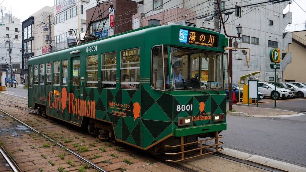 Green Hakodate City Tram Number 8001 on Route 5 to Yunokawa with Curry Ramai Advertising in Hokkaido, Japan Green Hakodate city tram number 8001 displaying route 5 to Yunokawa in Hokkaido, Japan, with orange Curry Ramai advertising, overhead electrical wires, tram tracks and buildings along the street.
