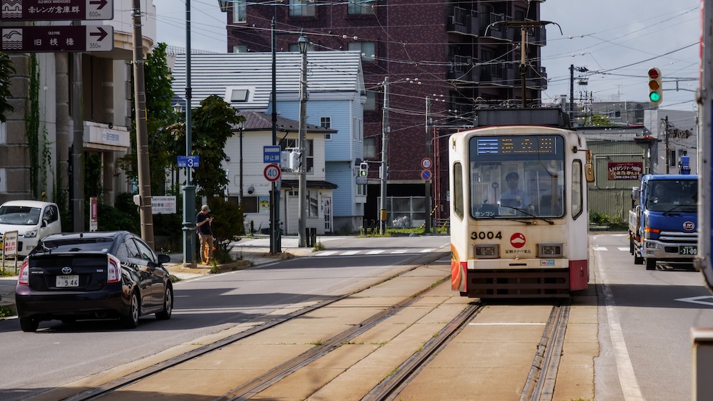 Hakodate City Tram travelling along street tracks in Hakodate, Hokkaido, Japan, with cars, overhead power lines and neighbourhood buildings surrounding the historic tram route.