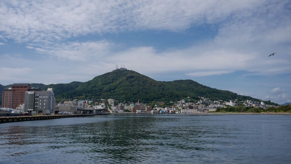 Scenic View Across Hakodate Harbour Toward Mt. Hakodate and the Waterfront City Skyline in Hokkaido, Japan View across Hakodate Harbour toward the forested slopes of Mt. Hakodate in Hokkaido, Japan, with waterfront buildings, a pier, calm blue water and a mountain summit topped with communications towers beneath a cloudy sky.