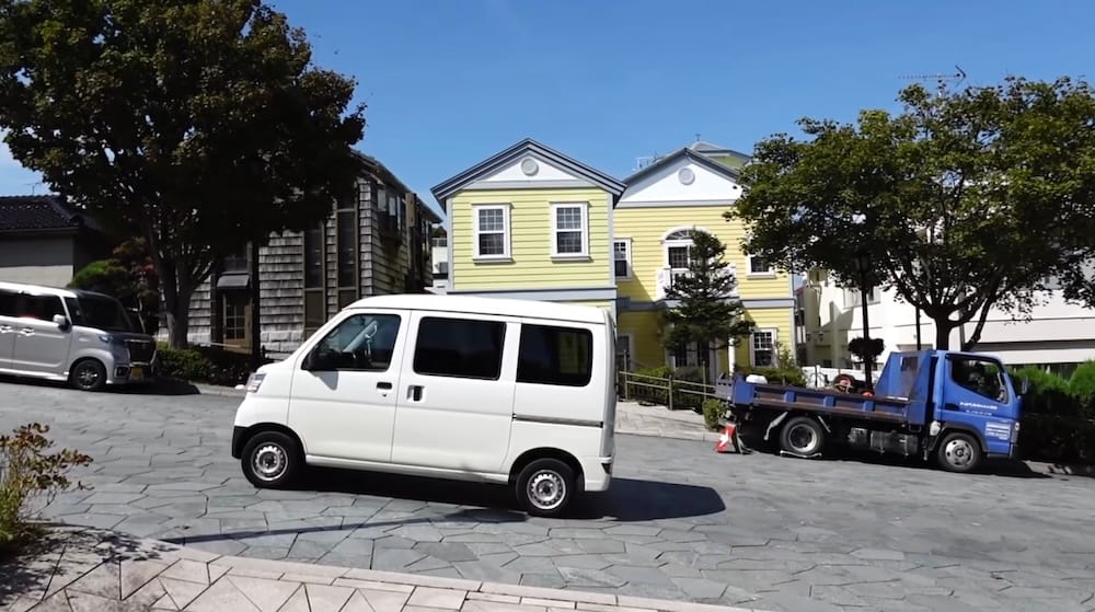 Auto pequeño subiendo una calle con pendiente en Hakodate, Hokkaido, Japón Un pequeño auto japonés blanco subiendo por una calle inclinada de Hakodate, Hokkaido, Japón, rodeado de edificios bajos, vehículos estacionados, árboles y cielo azul.