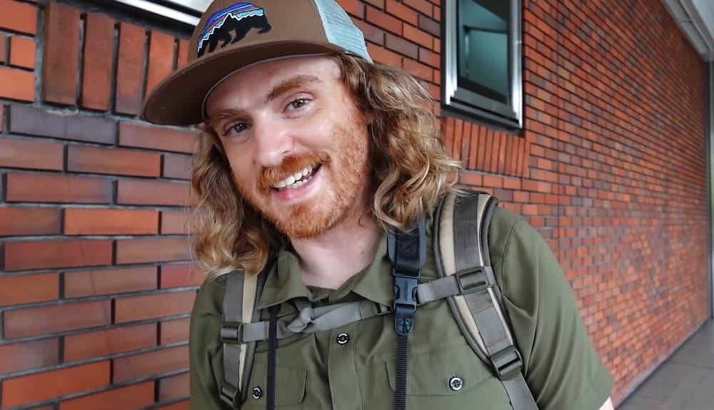 Samuel Jeffery sonríe mientras recorre Hakodate, Hokkaido, Japón, con camisa verde, gorra y mochila junto a una pared de ladrillos rojos durante un día de paseo por la ciudad.