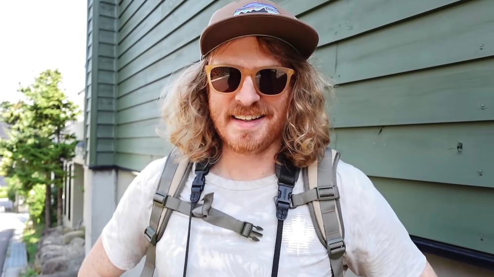 Samuel Jeffery smiles while sightseeing on foot in Hakodate, Hokkaido, Japan, wearing sunglasses, a cap and backpack beside a green-sided building on a sunny day.