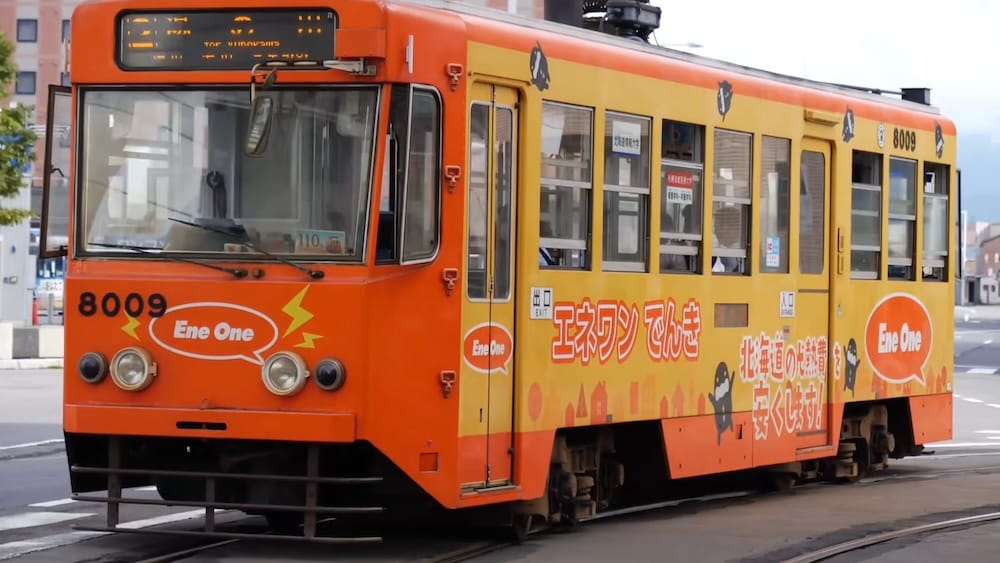 An orange and yellow Hakodate city tram numbered 8009, covered in Japanese and English advertising, runs on street tracks in Hakodate, Hokkaido, Japan, with buildings and a road behind it.