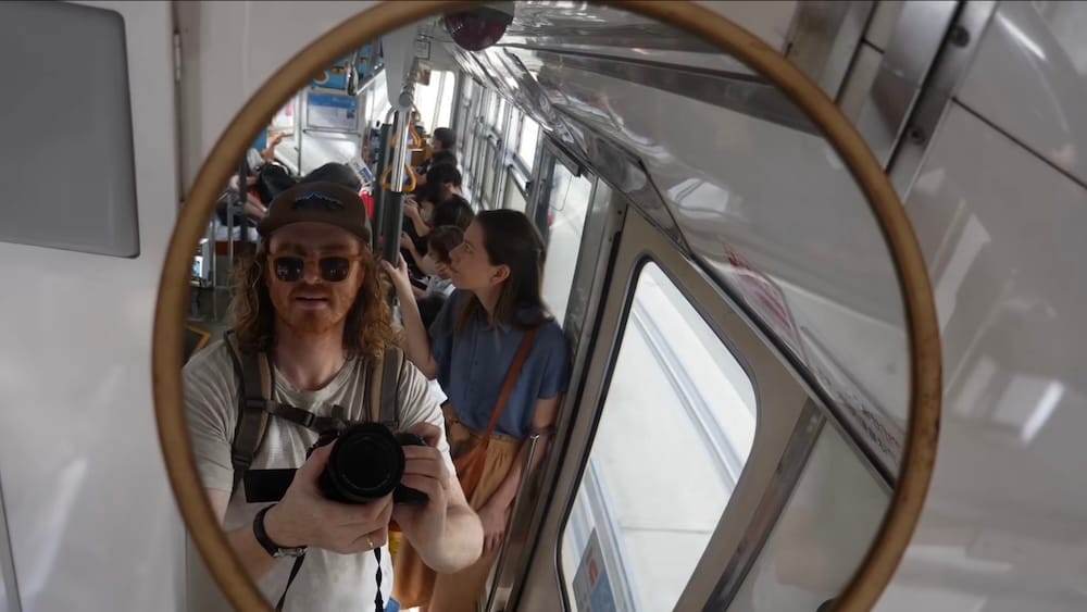 Samuel Jeffery, holding a camera, and Audrey Bergner are reflected in a round mirror inside a Hakodate city tram in Hokkaido, Japan, with other passengers seated and standing in the carriage behind them.