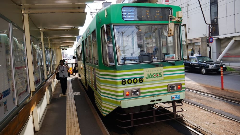 Tranvía urbano 8006 en un andén techado con piso podotáctil, tranvía de Hakodate, Hakodate, Hokkaido, Japón Un tranvía urbano de Hakodate a rayas verdes y blancas número 8006 espera en un andén techado con piso podotáctil en Hakodate, Hokkaido, Japón, con un pasajero en el andén y un taxi negro en la calle al lado.