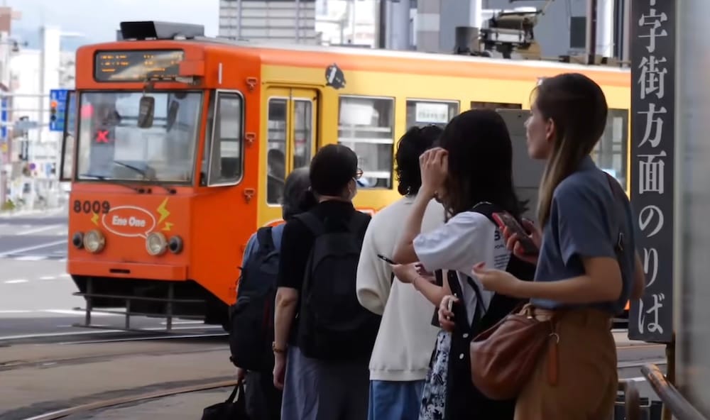 Audrey Bergner and other passengers wait at a tram platform in Hakodate, Hokkaido, Japan, as an orange and yellow city tram numbered 8009 approaches, beside a vertical Japanese platform sign.