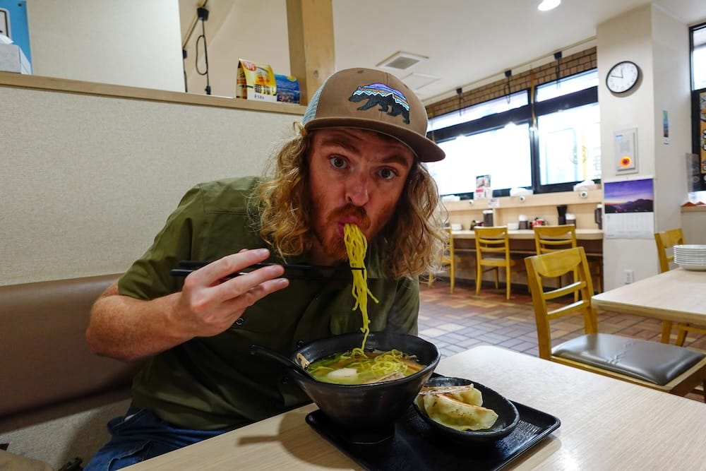 Samuel Jeffery slurps ramen noodles with chopsticks inside a casual restaurant in Hakodate, Hokkaido, Japan, with a bowl of noodles and a small plate of gyoza on the table.