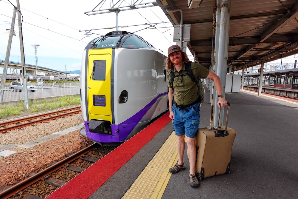Estación JR Hakodate, Hokkaido, Japón: Samuel Jeffery con su valija junto al tren Limited Express Hokuto Samuel Jeffery con una valija con ruedas y una mochila negra junto a un tren Limited Express Hokuto de JR Hokkaido, plateado, con frente amarillo, en el andén cubierto de la estación de Hakodate, Hokkaido, Japón.