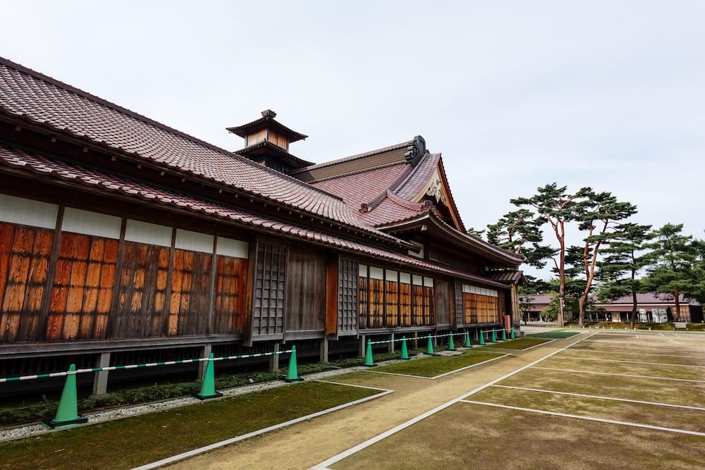 Exterior de la Oficina del Magistrado de Hakodate con techo de tejas y pinos, Parque Goryokaku, Hakodate, Hokkaido, Japón La Oficina del Magistrado de Hakodate en el Parque Goryokaku, Hokkaido, Japón: un edificio largo de madera con techo de tejas y paredes de listones de madera, conos verdes con una barrera de cuerda, líneas blancas en el suelo y pinos.