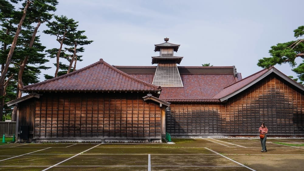 Audrey Bergner está parada en los terrenos de la Oficina del Magistrado de Hakodate en el Parque Goryokaku, Hokkaido, Japón, frente a sus largas paredes de madera y techos de tejas superpuestas, con pinos detrás y líneas blancas en el suelo.