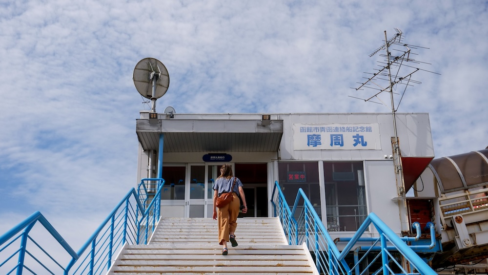 Audrey Bergner Climbing the Entrance Stairs to Mashu Maru Seikan Ferry Memorial Ship in Hakodate, Hokkaido, Japan Audrey Bergner climbs a wide outdoor staircase with bright blue railings toward the entrance of the Mashu Maru Seikan Ferry Memorial Ship in Hakodate, Hokkaido, Japan, beneath a cloudy sky and Japanese signage.