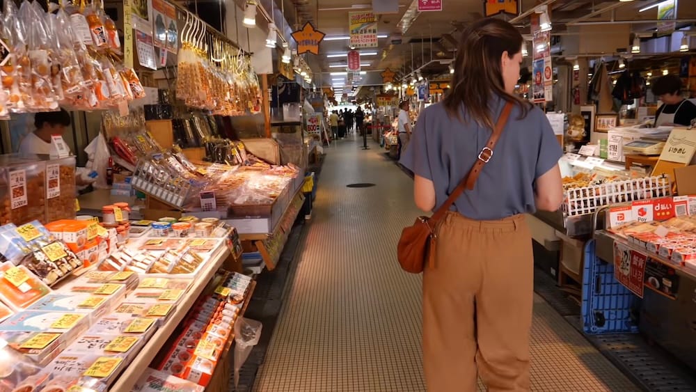 Walking the aisles of Hakodate Morning Market, Hakodate, Hokkaido, Japan – Audrey Bergner Audrey Bergner walks down a narrow tiled aisle at Hakodate Morning Market in Hakodate, Hokkaido, Japan, past stalls stacked with dried seafood, snacks and hanging goods under fluorescent lights.