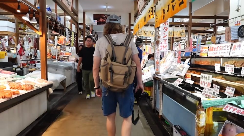 Samuel Jeffery walks through Hakodate Morning Market in Hakodate, Hokkaido, Japan, carrying a camera past seafood counters, hanging signs, packaged products and vendors inside the market aisle.