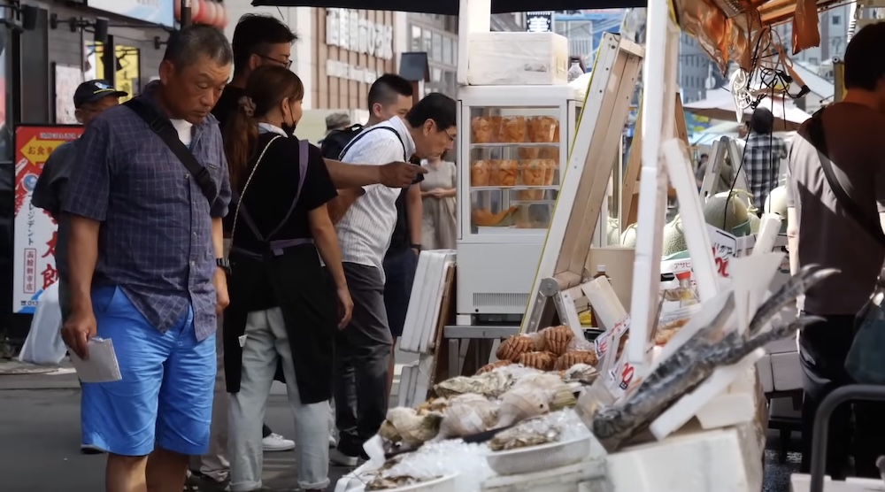 Actividad en Hakodate Morning Market con puestos de mariscos y compradores en Hakodate, Hokkaido, Japón Compradores pasan junto a puestos de mariscos frescos en Hakodate Morning Market, Hakodate, Hokkaido, Japón, donde se exhiben mariscos y otros productos en un pasillo exterior concurrido.