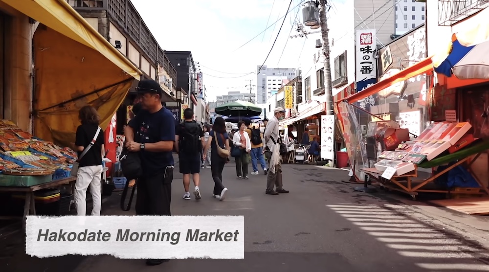 Shoppers walk between seafood stalls along a narrow lane at Hakodate Morning Market in Hakodate, Hokkaido, Japan, with colorful awnings, shop signs and fresh seafood displays on both sides.