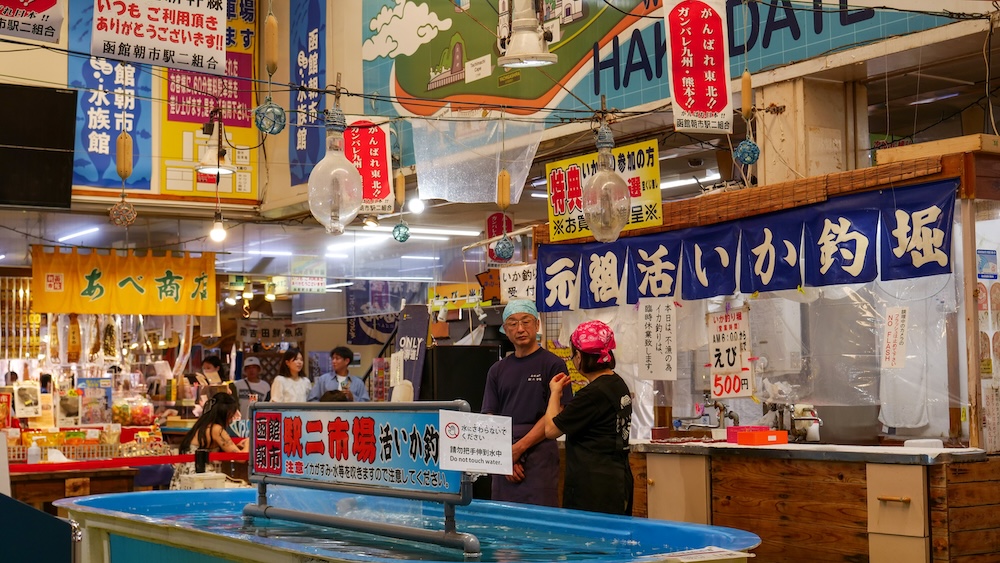 Tanque de pesca de calamares vivos dentro del Mercado Matutino de Hakodate, Hokkaido, Japón, con carteles japoneses azules, flotadores de vidrio colgantes, puestos de mariscos y dos personas junto al mostrador.