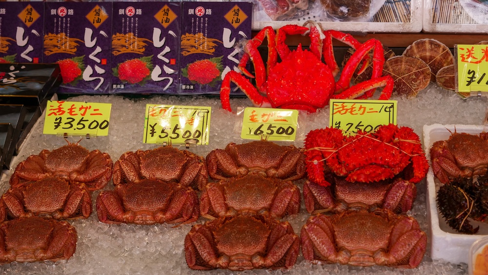 Fresh Hairy Crabs and Red King Crabs on Ice at Hakodate Morning Market in Hokkaido, Japan – Seafood Display Fresh brown hairy crabs and bright red king crabs displayed on crushed ice at Hakodate Morning Market in Hokkaido, Japan, with handwritten Japanese price signs and packaged salmon roe behind the seafood.