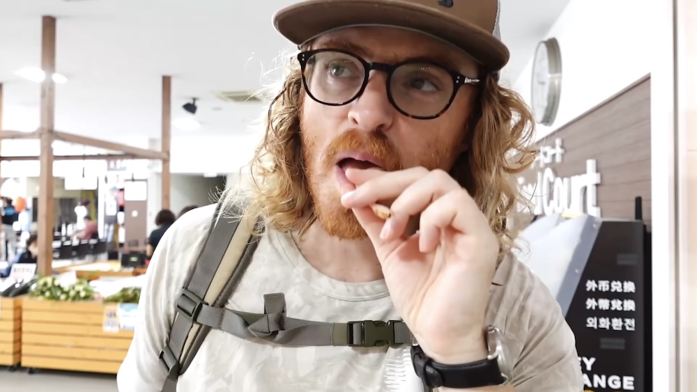 Samuel Jeffery con anteojos, gorra marrón y mochila mientras prueba un pequeño trozo de calamar dentro del Mercado Matutino de Hakodate, Hokkaido, Japón, con puestos y un mostrador de cambio de divisas detrás.