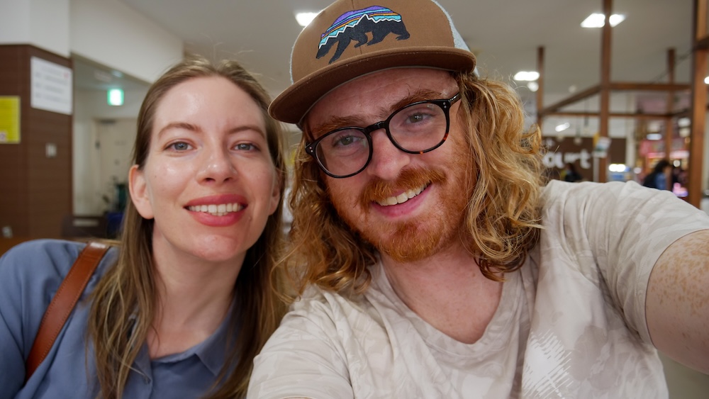 Audrey Bergner and Samuel Jeffery Visiting Hakodate Morning Market in Hokkaido, Japan – Travel Selfie Together Audrey Bergner and Samuel Jeffery smiling for a close-up selfie inside Hakodate Morning Market in Hokkaido, Japan, with Samuel wearing glasses and a brown baseball cap and indoor market lighting visible behind them.
