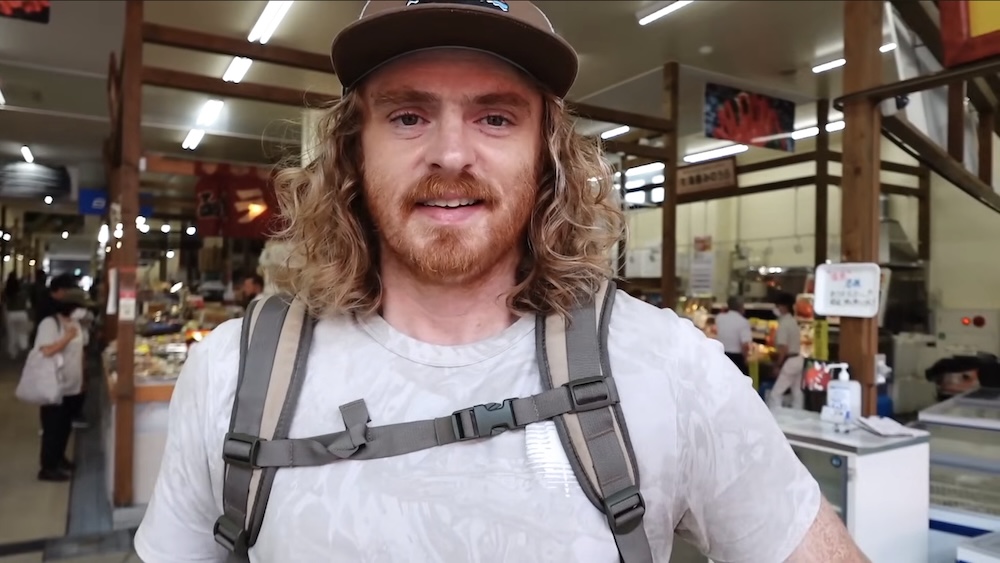 Samuel Jeffery Exploring Hakodate Morning Market in Hokkaido, Japan – Indoor Seafood Stalls and Market Visit Samuel Jeffery standing inside Hakodate Morning Market in Hokkaido, Japan, wearing a baseball cap, white T-shirt and grey travel backpack, with seafood counters, shoppers, wooden support beams and overhead lights behind him.