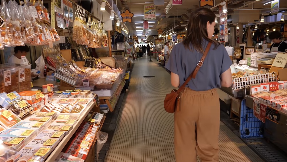 Audrey Bergner caminando por un pasillo interior del Mercado Matutino de Hakodate, Hokkaido, Japón, rodeada de puestos con mariscos envasados, productos secos colgantes, carteles de precios en japonés y otras mercaderías.