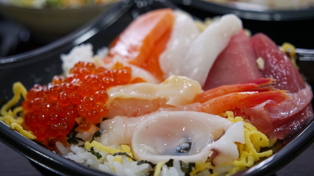 Desayuno de kaisendon con huevas de salmón, camarones, sashimi y moluscos en el Mercado Matutino de Hakodate, Hokkaido, Japón Primer plano de un kaisendon del Mercado Matutino de Hakodate, Hokkaido, Japón, servido en un recipiente negro con arroz, huevas de salmón, láminas de pescado crudo, camarones, moluscos y huevo en tiras.