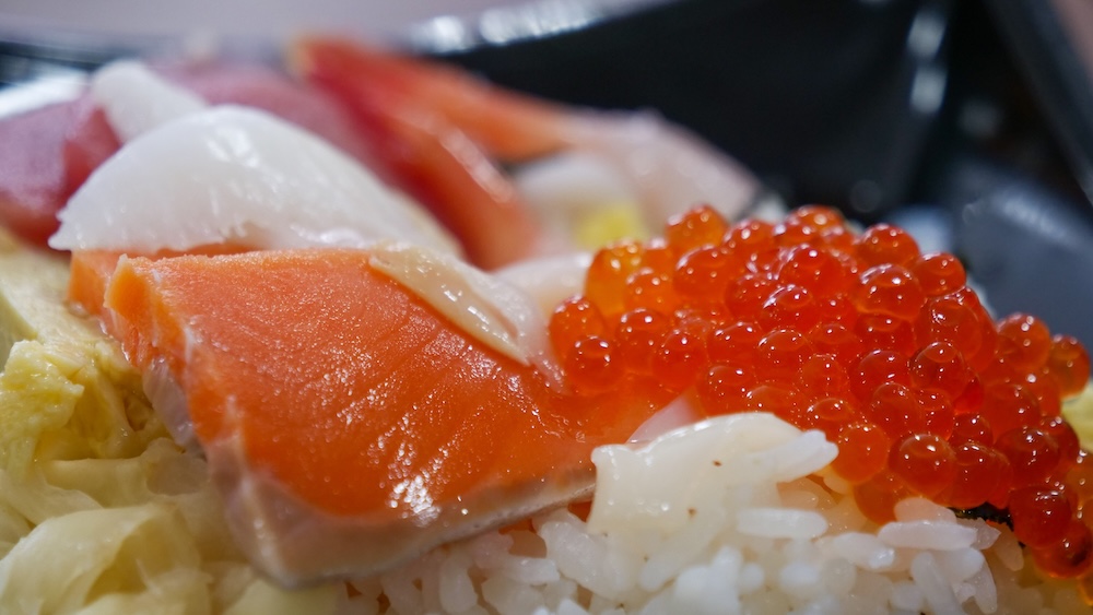 Primerísimo plano de un bol de arroz con mariscos kaisendon en el Mercado Matutino de Hakodate en Hokkaido, Japón, con gruesas fetas de sashimi de salmón naranja, huevas de salmón brillantes, pescado blanco y arroz al vapor con huevo amarillo.