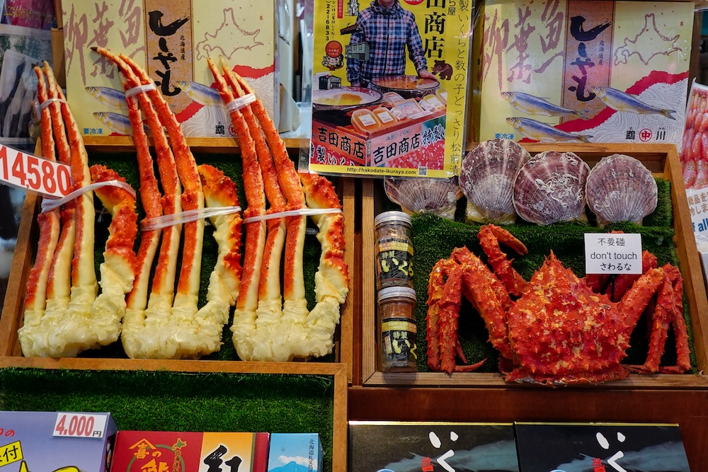 Puesto de mariscos en el Mercado Matutino de Hakodate, Hokkaido, Japón, con enormes patas de cangrejo real de color rojizo, un cangrejo entero de caparazón espinoso, conchas de vieiras, carteles publicitarios japoneses y precios.