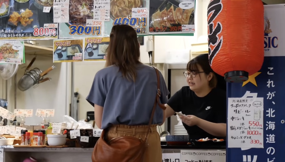 Audrey Bergner pidiendo un desayuno de mariscos en un mostrador con menús japoneses del Mercado Matutino de Hakodate, Hokkaido, Japón Audrey Bergner haciendo un pedido de desayuno en un mostrador del Mercado Matutino de Hakodate, Hokkaido, Japón, mientras habla con una empleada con anteojos bajo fotos de menús japoneses y un gran farol rojo.