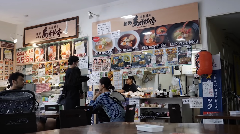 Interior del patio de comidas del Mercado Matutino de Hakodate en Hakodate, Hokkaido, Japón, con mostradores de restaurantes de mariscos y ramen cubiertos de menús japoneses con fotos, exhibidores coloridos, comensales sentados y personal preparando pedidos.