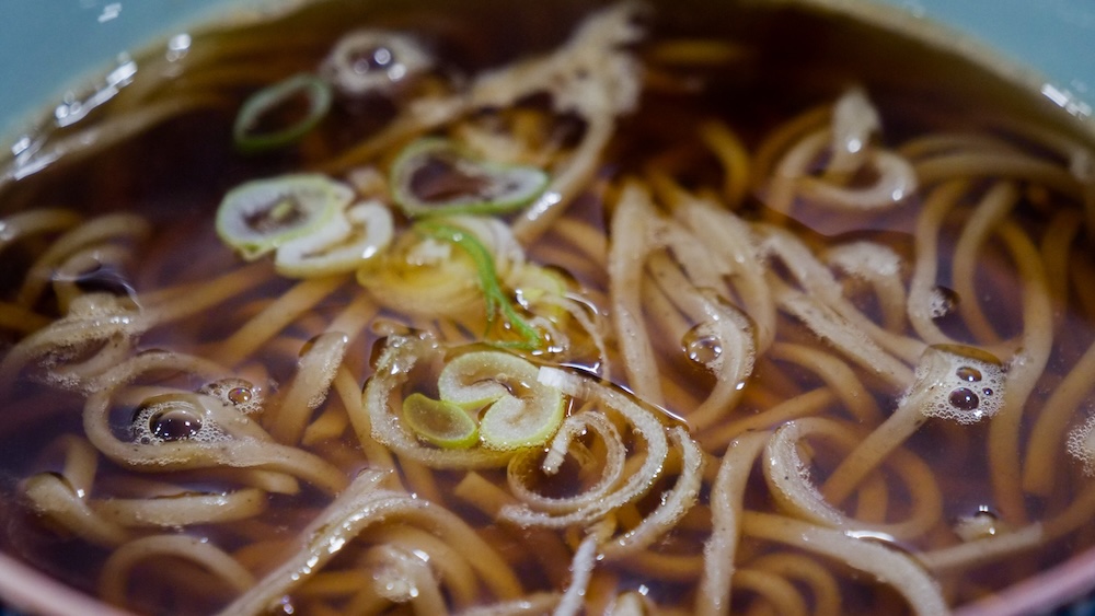 Sopa caliente de fideos soba con caldo oscuro y cebolla de verdeo en el Mercado Matutino de Hakodate, Hokkaido, Japón Primer plano de un bol de fideos soba en caldo oscuro en el Mercado Matutino de Hakodate, Hokkaido, Japón, con fideos marrones largos, rodajas de cebolla de verdeo, pequeñas burbujas y tiras finas de guarnición flotando.