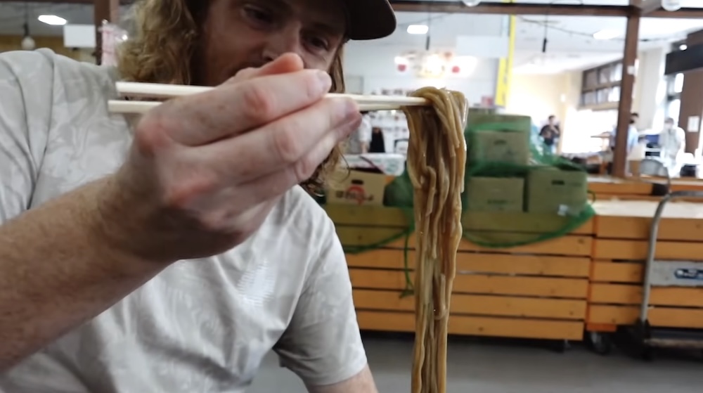 Samuel Jeffery levantando fideos soba calientes con palitos durante un desayuno de mariscos en el Mercado Matutino de Hakodate, Japón Samuel Jeffery levantando una porción larga de fideos marrones con palitos de madera durante el desayuno en el Mercado Matutino de Hakodate, Hokkaido, Japón, con mostradores y puestos de madera al fondo.