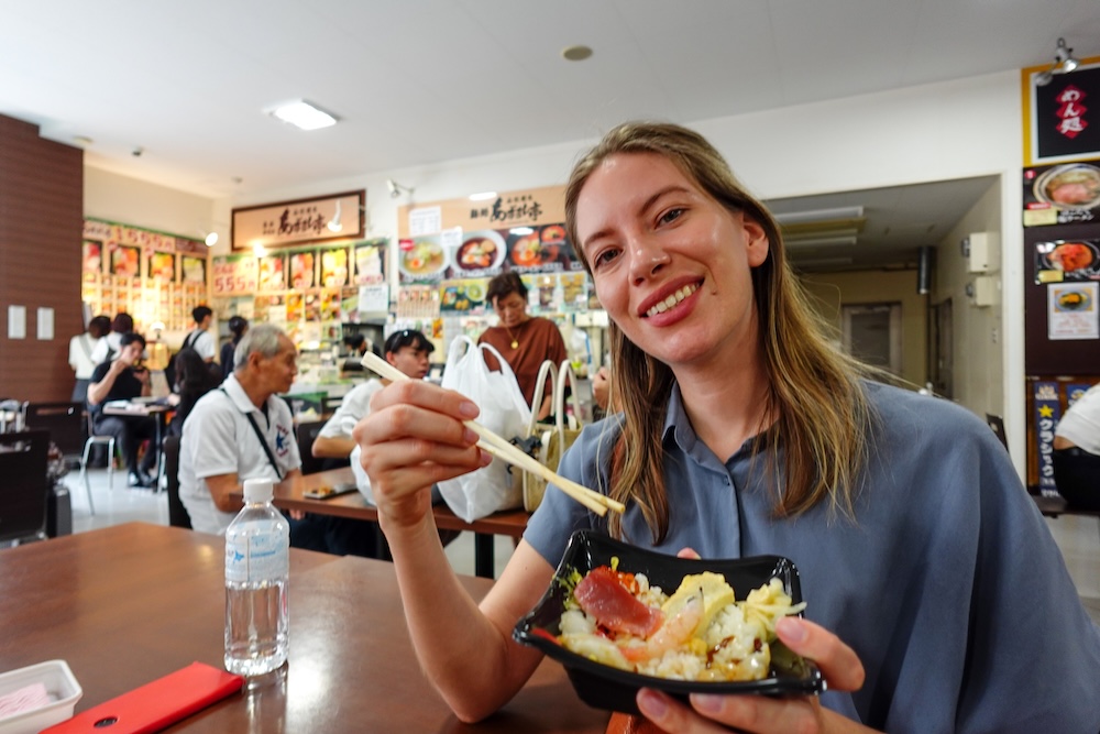 Seafood Rice Bowl Breakfast at Hakodate Morning Market, Hokkaido, Japan, with Audrey Bergner Audrey Bergner holds a seafood rice bowl and chopsticks while enjoying breakfast at Hakodate Morning Market in Hokkaido, Japan, with colourful Japanese restaurant menus and other diners in the background.