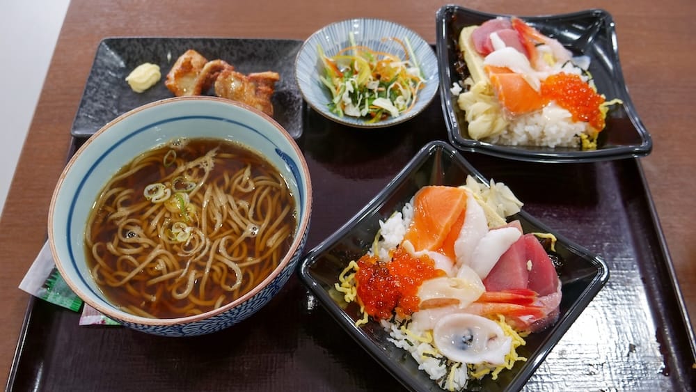 A Hakodate Morning Market meal in Hokkaido, Japan, with two seafood rice bowls topped with salmon, tuna, roe and other sashimi, alongside a bowl of noodles, gyoza and salad.