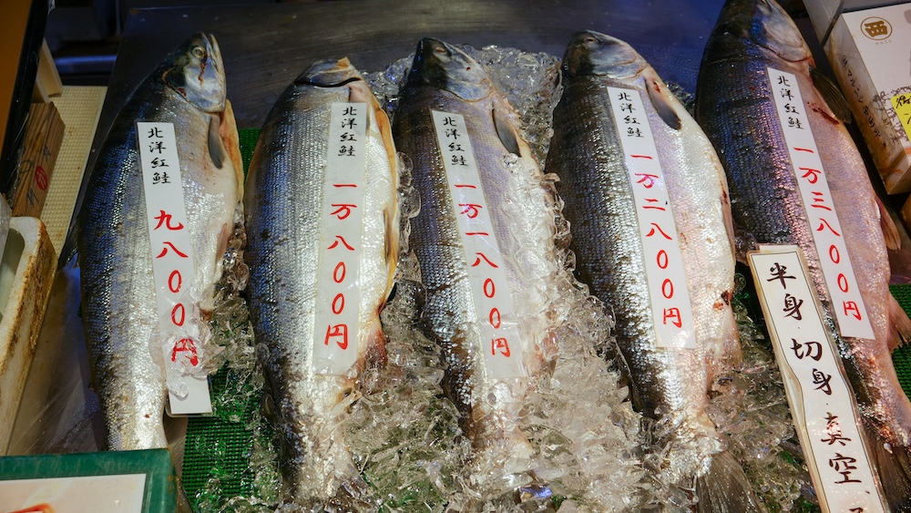 Cinco salmones enteros de gran tamaño exhibidos sobre hielo picado en el Mercado Matutino de Hakodate, Hokkaido, Japón, con etiquetas verticales de precios en japonés y un cartel que ofrece servicio de corte del pescado.