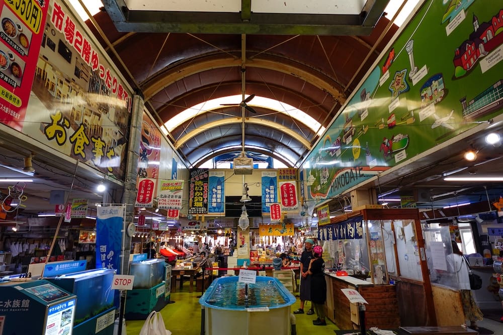 Vendors stand beside a live fish tank under the arched skylit roof of Hakodate Morning Market in Hakodate, Hokkaido, Japan, with hanging Japanese signs, seafood stalls and an illustrated Hakodate map mural.