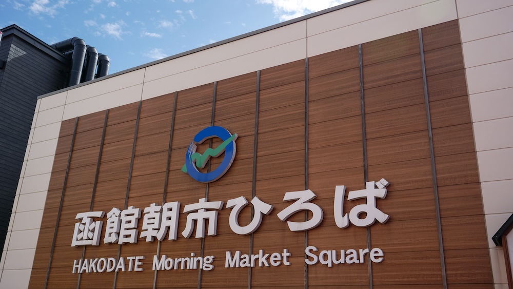 Exterior del edificio Hakodate Morning Market Square, conocido como Asaichi Hiroba, en Hakodate, Hokkaido, Japón, con grandes letras japonesas e inglesas, un logotipo de colores y una fachada revestida de madera bajo el cielo azul.