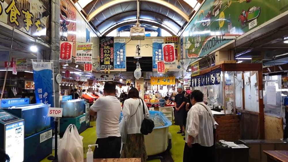 Shoppers browse seafood stalls under an arched roof at Hakodate Morning Market Square in Hakodate, Hokkaido, Japan, past live fish tanks, hanging Japanese signs and vendors behind the counters.