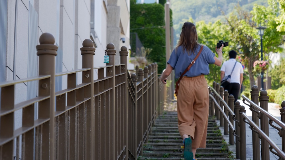 Audrey Bergner Climbing a Hillside Stairway in Motomachi Hakodate Hokkaido Japan During a Walking Tour Audrey Bergner climbing a stone stairway in Hakodate, Hokkaido, Japan, holding a camera beside a white building, with metal railings, leafy trees and another pedestrian farther up the steps.